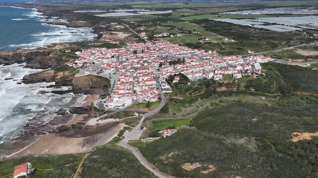 Aerial push-in drone shot of the picturesque coastal village Zambujeira do Mar with white houses and red roofs nestled on rugged cliffs by the Atlantic Ocean in Portugal. Part of the Rota Vicentina.
