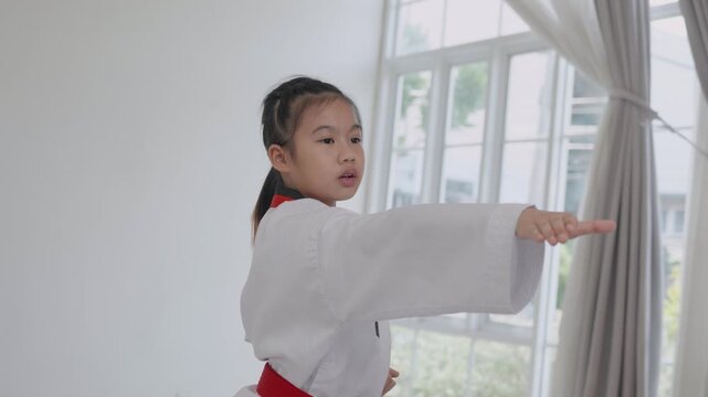 A child shows karate moves in a training room with white walls. The child wears a white uniform with a red belt. The setting is bright and simple, focusing on the training.