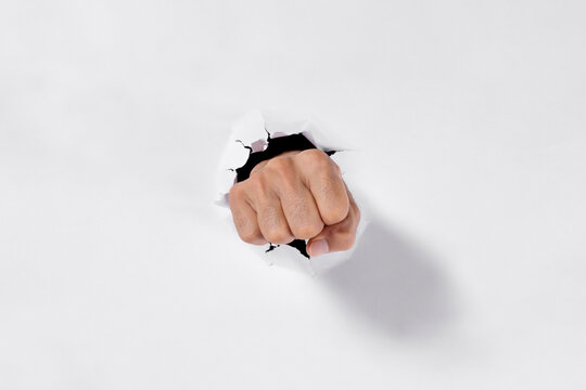 Man's clenched fist breaking through a torn white paper hole, strength, determination and breakthrough concept.