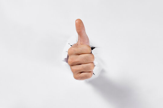 Thumbs up hand breaking through torn white paper with ripped hole on white background.