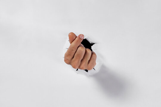 Male hand making a mini heart gesture through a torn hole in white paper, symbol of love and affection.