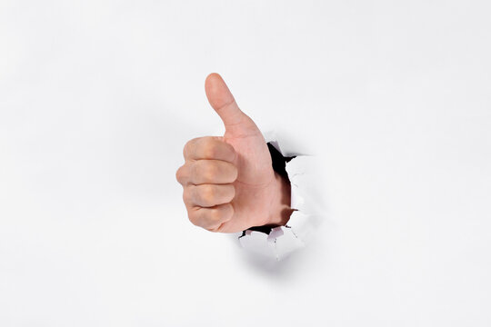 Man's hand giving a thumbs up through a torn white paper hole, approval and success concept.