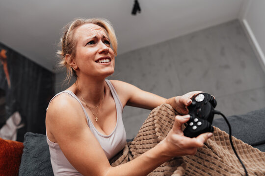 A cheerful female gamer relaxes under a warm blanket while holding a gamepad. She enjoys a fun gaming session on a comfortable grey sofa with snacks nearby.