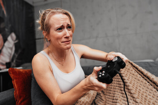 An intense female gamer reacts with a passionate grimace while holding a black controller under a cozy blanket on the sofa.