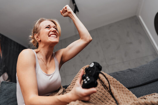 A cheerful female gamer laughs and raises her fist in triumph while holding a controller on her living room sofa.