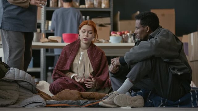 Young adult Caucasian woman and African American man sitting in shelter having conversation, receiving water bottle from volunteer. Perfect for charity campaigns and humanitarian aid promotions
