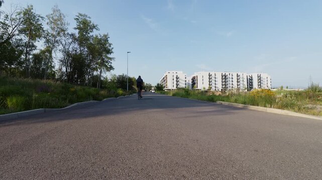 Rear view of a man with a backpack riding a bicycle on an asphalt path heading towards a modern residential area