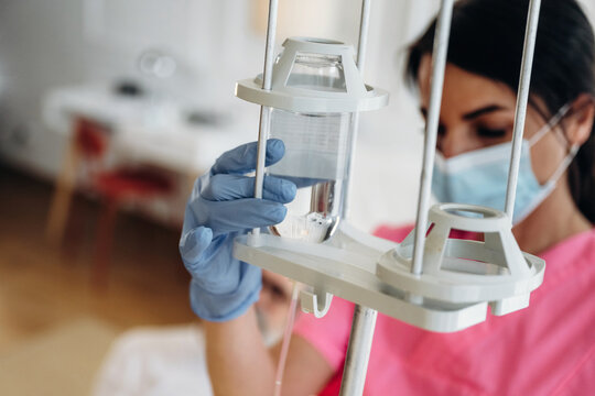 Female nurse in pink scrubs adjusts IV drip with clear fluid in hospital room, medical equipment and patient bed visible in the background