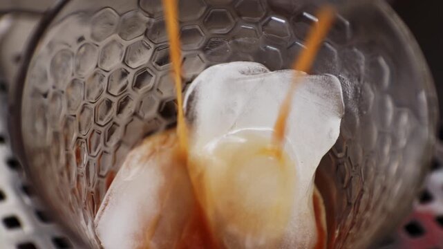 Close up overhead view of hot espresso pouring from a coffee machine over a large ice cube in a glass