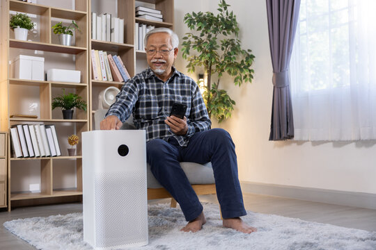 Senior man air purifier smartphone living room, calm male adjusting home appliance near shelf, plant, window, curtain