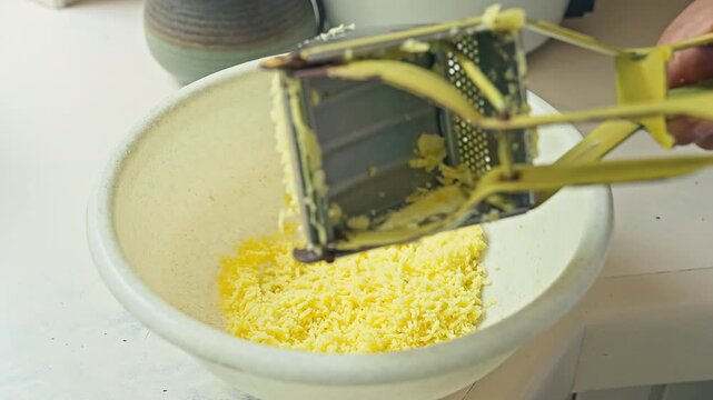 A metal potato ricer to mash yellow potatoes in a white bowl
