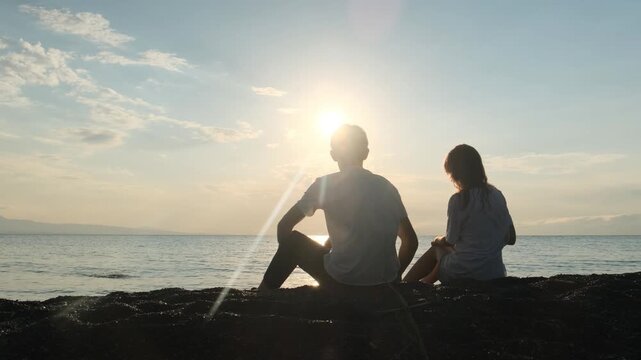 Two happy teenagers, a boy and a girl, sit together on the seashore at sunset, throwing stones into the water. Quiet nature, heartfelt conversation, first love, friendship, romantic couple