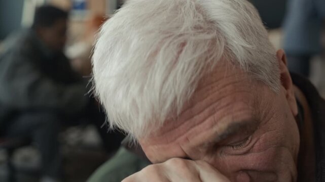 Caucasian senior man taking off glasses and rubbing eyes, sitting in humanitarian aid shelter. Clip is perfect for charity campaigns, social support services, and mental health awareness videos