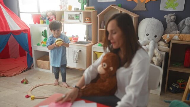 Woman holds a plush monkey and gestures with hands as little boy plays at toy kitchen in preschool building; calm caregiving.