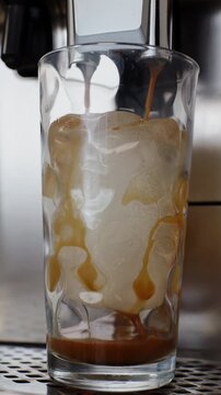 Vertical front view of hot espresso pouring from a coffee maker into a tall glass filled with large ice cubes