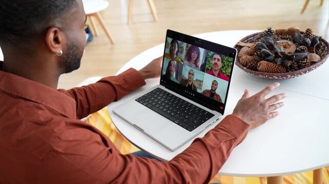 Over the shoulder view of a black man sitting at a table participating in an online group therapy session on his laptop, listening and talking with other participants in a video conference call