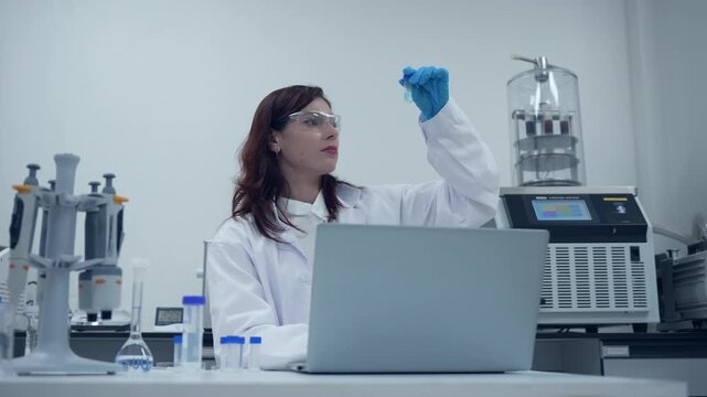 Female scientist typing on a laptop computer and examining a test tube sample in a modern laboratory research facility