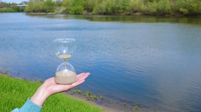 Hourglass in nature. Person's hand holds a glass hourglass over a tranquil river, the sand flowing from the top bulb to the bottom, symbolizing the inexorable passage of time and the beauty of nature
