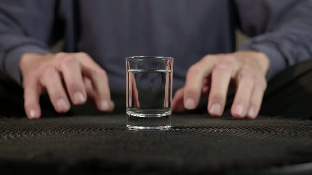 Male hands nervously touching shot glass of alcohol. Anxious man sitting at a dark table, his nervous hands restlessly moving around a shot glass full of clear liquor, contemplating whether to drink
