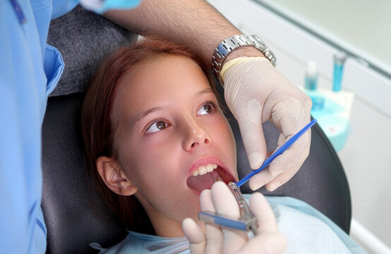Close up over the shoulder shot of a dentist using a dental handpiece drill and mirror to treat caries or polish teeth of a brave young girl patient sitting in a dental chair.