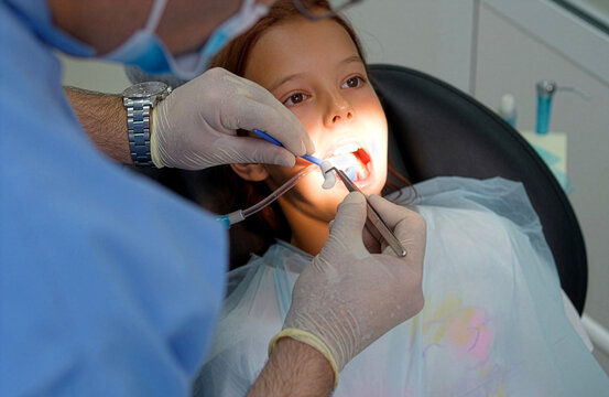 Close up high angle shot of a professional dentist in medical gloves using a dental mirror and probe tool to examine teeth of a young girl patient lying in a black dental chair at a clinic.
