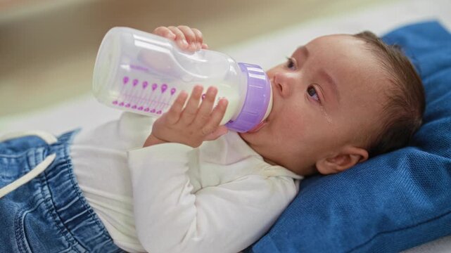 Baby boy feeding from milk bottle, tiny hand grasping teat while lying on pillow in building; nurturing comfort bonding.