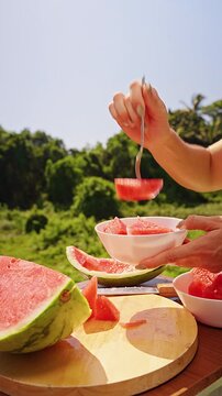 Vertical video. Woman Picking Up Fresh Watermelon Pieces With a Fork Into a Bowl Outdoors, Close Up, Healthy Hydration, Fresh Summer Eating