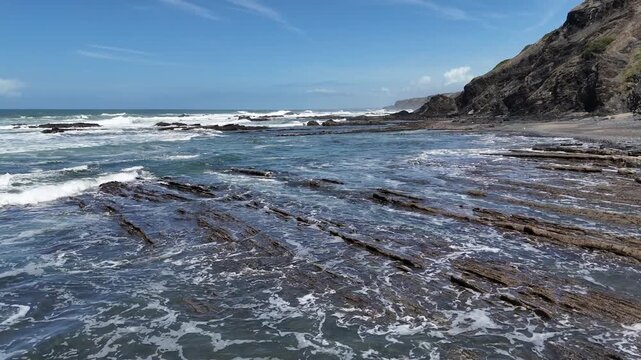 An aerial drone push-in shot captures the stunning, remote coastline of Portugal, part of the Rota Vicentina's Fisherman's Trail, with blue ocean waves crashing on rocky cliffs.