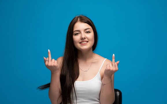 Young brunette woman smiling, gesturing middle fingers, expressing rebellion and disrespect on blue background
