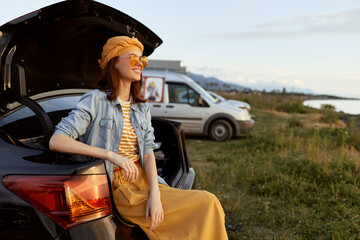 Naklejka premium Woman lifestyle outdoor casual summer travel, sits on car trunk in countryside, wearing denim dress, hat and sunglasses, smiling under bright sunshine, relaxed pose and peaceful mood