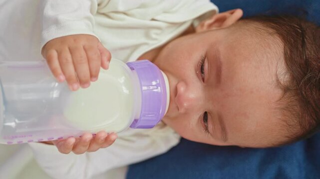 Hispanic baby boy lying on blue blanket holding milk bottle to mouth with tiny hand in home nursery; calm bonding.