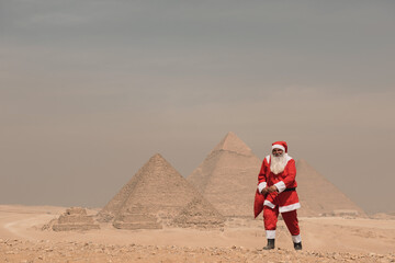 Fototapeta premium Iconic pyramid backdrop frames a surreal and festive Santa Claus in full red costume holding a gift bag on the sandy Giza plateau under a hazy