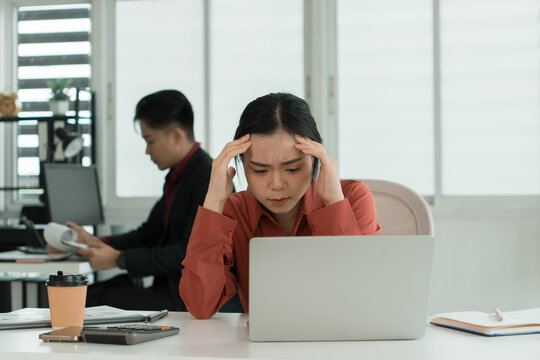 Stressed Businesswoman Working on Laptop with Colleague in Background