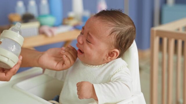 Hispanic baby boy crying with clenched fists and tear streaks, refusing a bottle while seated in a nursery highchair near crib and changing table; hunger distress.