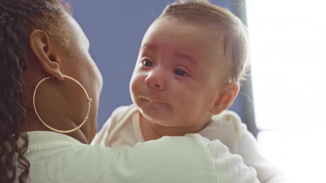 Baby boy face with milk and drool on mouth held on caregiver shoulder near bright window inside building with visible gold hoop earring; gentle comfort.