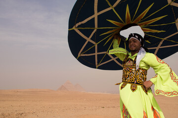 Fototapeta premium Proud Egyptian tanoura dancer in neon embroidered robe raises large spinning disc before ancient Giza Pyramids in overcast desert