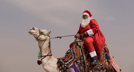 Fototapeta premium Humorous camel rider in full Santa Claus red suit riding a decorated dromedary under overcast desert sky bold multicultural holiday concept