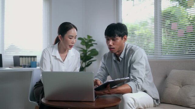Asian colleagues collaborating on project, discussing ideas with laptop and clipboard in a modern office living room setting