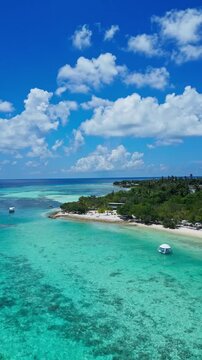 Aerial reef patterns along turquoise bay, meandering sandbars and shallow coral gardens, anchored boats near palmfringed shoreline, beachside huts and wide cloudscapes, Maldives, vertical video