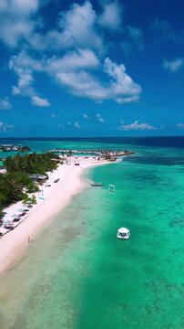 Aerial reef patterns along turquoise bay, meandering sandbars and shallow coral gardens, anchored boats near palmfringed shoreline, beachside huts and wide cloudscapes, Maldives, vertical video