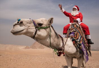 Fototapeta premium Festive Santa Claus riding a white camel and waving cheerfully with ancient Giza Pyramids rising through desert haze in background ideal for global