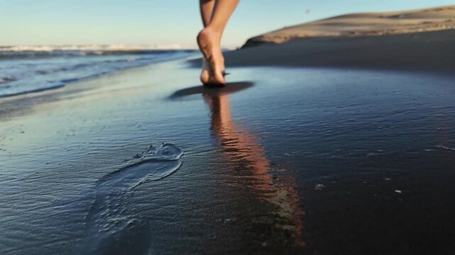 Low-angle shot of a person's bare feet walking on the wet sand of a beach at sunrise, leaving footprints behind as the morning light reflects on the glistening shoreline
