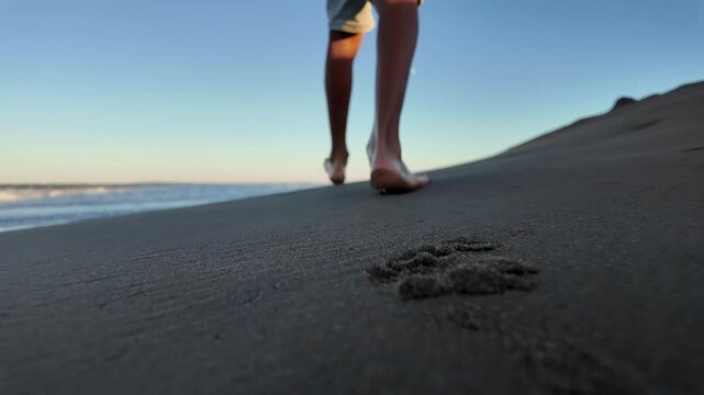 Person walking barefoot along a sandy beach during sunset, leaving footprints in the wet sand as the ocean waves gently roll in, with a focus on their feet and legs