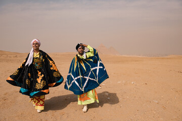 Fototapeta premium Two Egyptian whirling dancers wearing vivid embroidered traditional costumes strike theatrical poses on the Giza desert plateau with the iconic