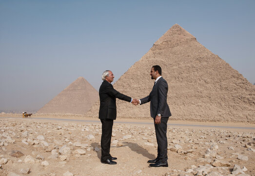 Business handshake between a younger Arab executive and senior Western professional at iconic Giza Pyramids desert plateau sealing a confident
