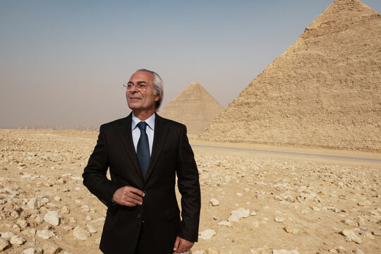 Giza Pyramids flank a solitary senior Arab businessman in dark suit gazing upward with composed gravitas ideal for executive leadership global
