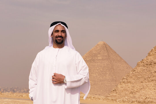 Confident gulf portrait of smiling emirati man in white kandura with luxury watch standing before ancient Giza Pyramids Egypt perfect for MENA