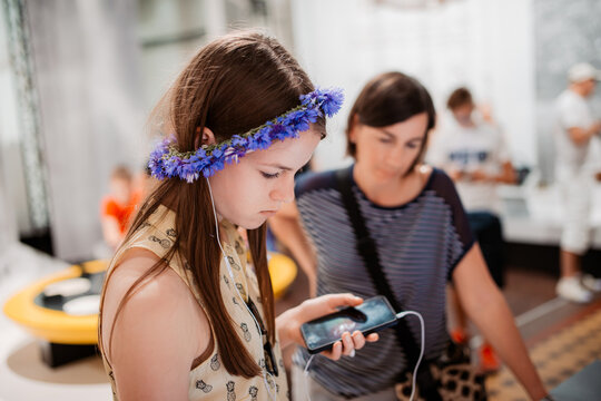 Profile View of a Focused Young Girl Wearing a Cornflower Crown and Earphones, Looking at a Smartphone inside an Interactive Science Museum Exhibit