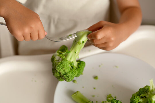 A child sits at a table and uses a fork to eat broccoli. The child appears focused on the food. The setting looks like a home kitchen