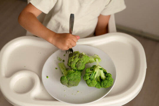 A child sits at a table in a high chair holding a fork and eating broccoli from a white plate. The setting is casual and simple